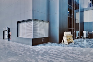 A modern building exterior with sleek, dark gray panels and large grid windows. A series of metal vents is visible on the left side, and outdoor air conditioning units are mounted on the wall. Wooden construction barriers and additional air conditioning units are seen at the base of the building.