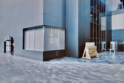 A modern building exterior with sleek, dark gray panels and large grid windows. A series of metal vents is visible on the left side, and outdoor air conditioning units are mounted on the wall. Wooden construction barriers and additional air conditioning units are seen at the base of the building.