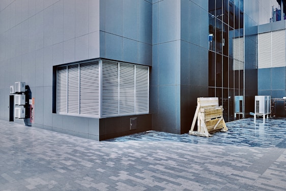 A modern building exterior with sleek, dark gray panels and large grid windows. A series of metal vents is visible on the left side, and outdoor air conditioning units are mounted on the wall. Wooden construction barriers and additional air conditioning units are seen at the base of the building.