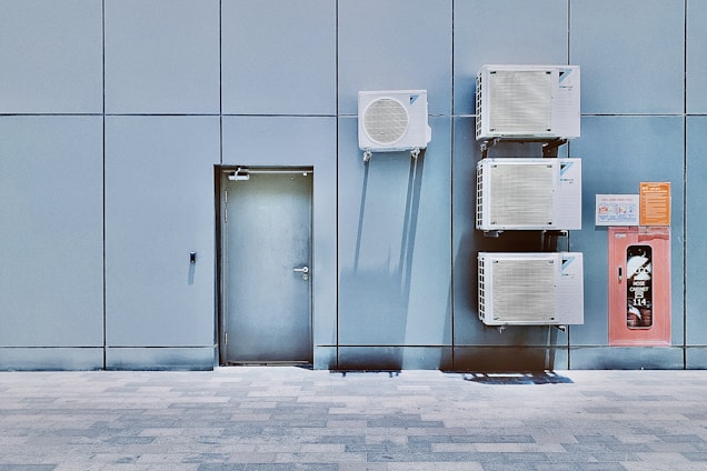 A blue tiled wall with a closed metal door and several air conditioning units mounted above and beside it. Next to the AC units is a red hose cabinet with a fire safety sign.