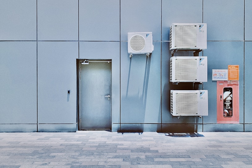 A blue tiled wall with a closed metal door and several air conditioning units mounted above and beside it. Next to the AC units is a red hose cabinet with a fire safety sign.