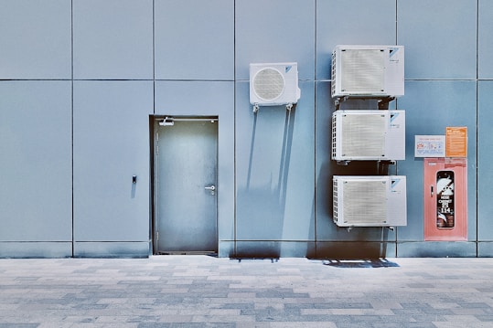 A blue tiled wall with a closed metal door and several air conditioning units mounted above and beside it. Next to the AC units is a red hose cabinet with a fire safety sign.