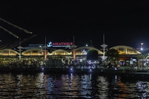 A night scene of a bustling riverfront destination, Asiatique, with illuminated arches and various signs in neon and warm lights. The water reflects the bright lights from the buildings and signage, adding to the lively atmosphere. People can be seen walking along the waterfront, enjoying the vibrant nightlife.