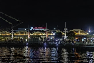 A night scene of a bustling riverfront destination, Asiatique, with illuminated arches and various signs in neon and warm lights. The water reflects the bright lights from the buildings and signage, adding to the lively atmosphere. People can be seen walking along the waterfront, enjoying the vibrant nightlife.