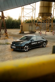 A modern black car with the label 'IONIQ 6' is parked in an industrial setting. Surrounding the vehicle are metal structures, storage silos, and piles of gravel. The area is open and quiet, with a mix of natural and industrial elements.