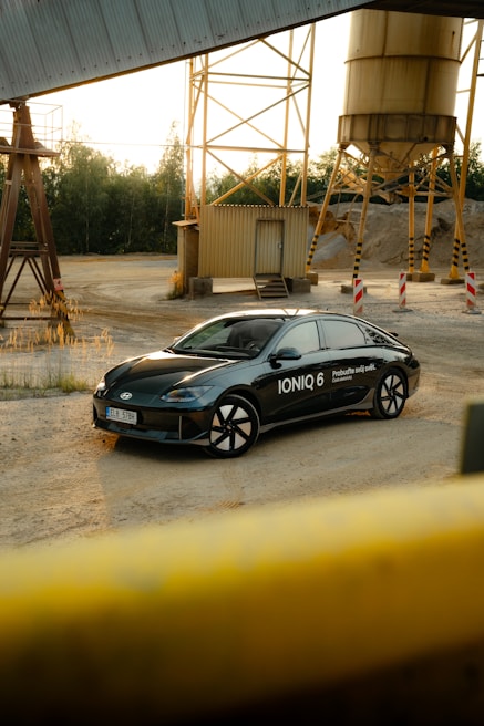 A modern black car with the label 'IONIQ 6' is parked in an industrial setting. Surrounding the vehicle are metal structures, storage silos, and piles of gravel. The area is open and quiet, with a mix of natural and industrial elements.