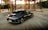 A sleek black Hyundai Ioniq 6 electric car is parked in an industrial area with metal structures and sandy terrain in the background. The sun is setting, casting a warm glow over the scene. The car features sharp, modern design lines and displays branding details on its side.