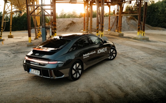 A sleek black Hyundai Ioniq 6 electric car is parked in an industrial area with metal structures and sandy terrain in the background. The sun is setting, casting a warm glow over the scene. The car features sharp, modern design lines and displays branding details on its side.