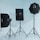 Three photography light stands equipped with softboxes are arranged in a studio setting. The softboxes vary in shape, with two rectangular and one octagonal. The stands are set against a plain white background, reflecting a minimalist studio environment.