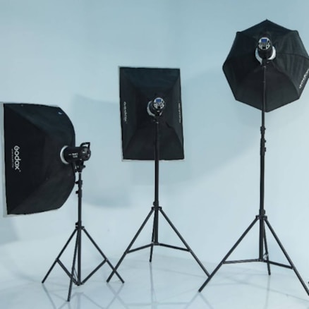 Three photography light stands equipped with softboxes are arranged in a studio setting. The softboxes vary in shape, with two rectangular and one octagonal. The stands are set against a plain white background, reflecting a minimalist studio environment.
