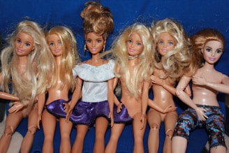 A group of six fashion dolls with detailed hair and various outfits is arranged in a row. Most of the dolls have long blonde hair, except for one with darker hair tied in an updo. Some of the dolls are wearing minimal clothing while one is dressed in a white top and purple skirt, and another in colorful leggings.