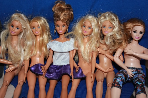 A group of six fashion dolls with detailed hair and various outfits is arranged in a row. Most of the dolls have long blonde hair, except for one with darker hair tied in an updo. Some of the dolls are wearing minimal clothing while one is dressed in a white top and purple skirt, and another in colorful leggings.