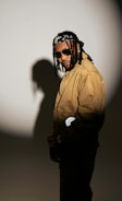 A person wearing sunglasses and a brown jacket stands against a spotlight that casts their shadow on a plain background. They have long braided hair tied with a patterned bandana and are looking towards the camera with a subtle smile.