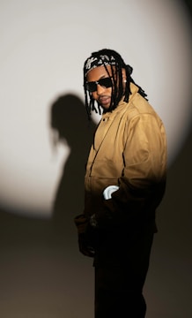 A person wearing sunglasses and a brown jacket stands against a spotlight that casts their shadow on a plain background. They have long braided hair tied with a patterned bandana and are looking towards the camera with a subtle smile.
