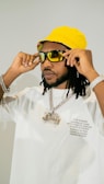 A person wearing a bright yellow bucket hat and large sunglasses, adjusting the glasses with both hands. The individual is dressed in a white shirt with printed text on the front and is wearing a chain necklace with a large pendant. The overall appearance is stylish and fashion-forward, with accessories that emphasize a bold aesthetic.