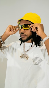 A person wearing a bright yellow bucket hat and large sunglasses, adjusting the glasses with both hands. The individual is dressed in a white shirt with printed text on the front and is wearing a chain necklace with a large pendant. The overall appearance is stylish and fashion-forward, with accessories that emphasize a bold aesthetic.
