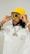 A person wearing a bright yellow bucket hat and large sunglasses, adjusting the glasses with both hands. The individual is dressed in a white shirt with printed text on the front and is wearing a chain necklace with a large pendant. The overall appearance is stylish and fashion-forward, with accessories that emphasize a bold aesthetic.