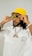 A person wearing a bright yellow bucket hat and large sunglasses, adjusting the glasses with both hands. The individual is dressed in a white shirt with printed text on the front and is wearing a chain necklace with a large pendant. The overall appearance is stylish and fashion-forward, with accessories that emphasize a bold aesthetic.