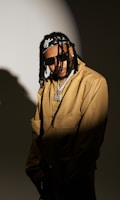 A person wearing sunglasses and a beige jacket, accessorized with a chain necklace and a bandana over braided hair, stands against a dark, moody background with a spotlight effect.