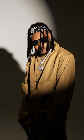 A person wearing sunglasses and a beige jacket, accessorized with a chain necklace and a bandana over braided hair, stands against a dark, moody background with a spotlight effect.