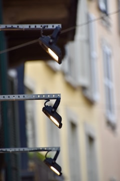 Three outdoor spotlights are mounted on a metal bracket against a building facade. The background is blurred with pastel-colored walls and windows, providing a soft contrast to the industrial lighting fixtures.
