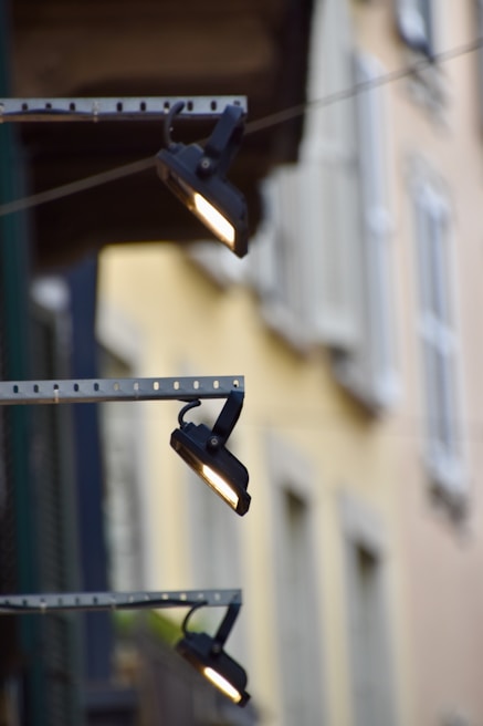 Three outdoor spotlights are mounted on a metal bracket against a building facade. The background is blurred with pastel-colored walls and windows, providing a soft contrast to the industrial lighting fixtures.