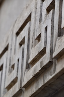 A close-up view of a stone wall featuring intricate geometric patterns carved into the surface. The design appears three-dimensional, with shadows emphasizing the depth of the carvings. The stone texture is rough and weathered, adding an element of aged character.