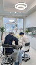 Two dental professionals are working on a patient seated in a dental chair within a modern dental office. The room has white walls, a large window, and a ceiling light fixture with circular designs. Dental equipment and tools are visible around the chair, and a potted plant adds greenery to the space.