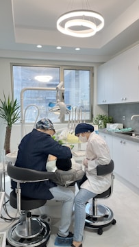Two dental professionals are working on a patient seated in a dental chair within a modern dental office. The room has white walls, a large window, and a ceiling light fixture with circular designs. Dental equipment and tools are visible around the chair, and a potted plant adds greenery to the space.