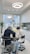Two dental professionals are working on a patient seated in a dental chair within a modern dental office. The room has white walls, a large window, and a ceiling light fixture with circular designs. Dental equipment and tools are visible around the chair, and a potted plant adds greenery to the space.