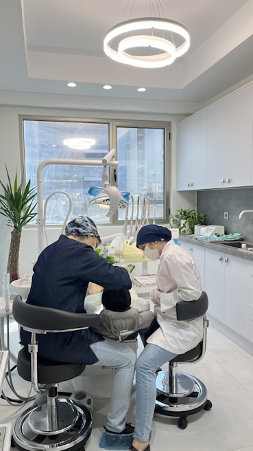 Two dental professionals are working on a patient seated in a dental chair within a modern dental office. The room has white walls, a large window, and a ceiling light fixture with circular designs. Dental equipment and tools are visible around the chair, and a potted plant adds greenery to the space.