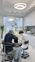 Two dental professionals are working on a patient seated in a dental chair within a modern dental office. The room has white walls, a large window, and a ceiling light fixture with circular designs. Dental equipment and tools are visible around the chair, and a potted plant adds greenery to the space.