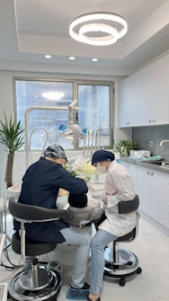 Two dental professionals are working on a patient seated in a dental chair within a modern dental office. The room has white walls, a large window, and a ceiling light fixture with circular designs. Dental equipment and tools are visible around the chair, and a potted plant adds greenery to the space.
