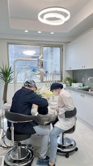 Two dental professionals are working on a patient seated in a dental chair within a modern dental office. The room has white walls, a large window, and a ceiling light fixture with circular designs. Dental equipment and tools are visible around the chair, and a potted plant adds greenery to the space.