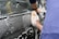 A person is scrubbing the side of a car with a sponge covered in soapy water, creating white suds on the black vehicle. The forearms and hands of the person are visible, wearing a blue sleeve.