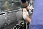 A person is scrubbing the side of a car with a sponge covered in soapy water, creating white suds on the black vehicle. The forearms and hands of the person are visible, wearing a blue sleeve.