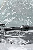 A windshield covered in soapy water and foam, with a windshield wiper visible in the image.