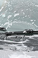 A windshield covered in soapy water and foam, with a windshield wiper visible in the image.