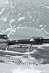 A windshield covered in soapy water and foam, with a windshield wiper visible in the image.