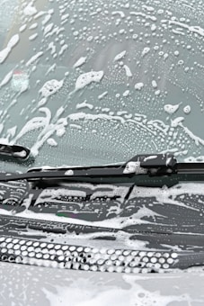 A windshield covered in soapy water and foam, with a windshield wiper visible in the image.