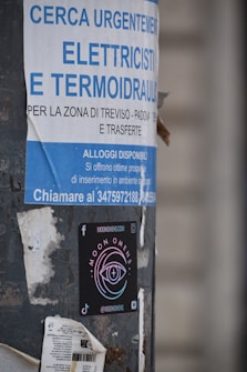 A pole displays a large advertisement for electricians and plumbers with urgent hiring needs in Italian. Below this is a colorful sticker for 'Moon Omens' featuring a stylized eye logo, alongside social media icons and web address.
