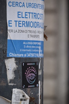 A pole displays a large advertisement for electricians and plumbers with urgent hiring needs in Italian. Below this is a colorful sticker for 'Moon Omens' featuring a stylized eye logo, alongside social media icons and web address.