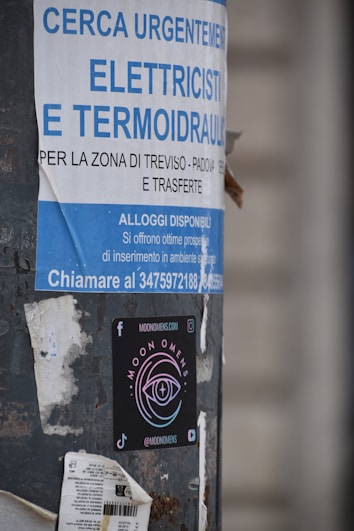 A pole displays a large advertisement for electricians and plumbers with urgent hiring needs in Italian. Below this is a colorful sticker for 'Moon Omens' featuring a stylized eye logo, alongside social media icons and web address.