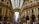 A bustling shopping gallery features ornate architectural details with arches and glass ceilings, while numerous people stroll along the corridor. Designer brand stores like Dior and Prada are visible on the ground floor.