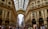 A bustling shopping gallery features ornate architectural details with arches and glass ceilings, while numerous people stroll along the corridor. Designer brand stores like Dior and Prada are visible on the ground floor.