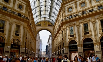 A bustling shopping gallery features ornate architectural details with arches and glass ceilings, while numerous people stroll along the corridor. Designer brand stores like Dior and Prada are visible on the ground floor.