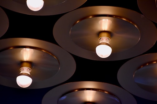 A series of round, metallic ceiling fixtures with exposed light bulbs. The fixtures are arranged in a repetitive, geometric pattern against a dark background, casting a soft, warm glow.
