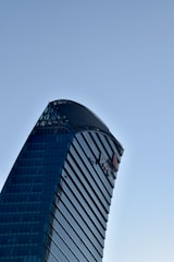 A modern skyscraper with a unique curved design and glass facade against a clear blue sky. The building features prominent logos and sleek architectural lines.