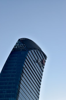 A modern skyscraper with a unique curved design and glass facade against a clear blue sky. The building features prominent logos and sleek architectural lines.