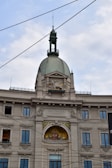 A historic European building with ornate architectural details, including a prominent dome with a decorative spire and windows. The facade features intricate stone carvings and a colorful fresco or mosaic beneath the central arch. Several windows are symmetrically placed, and the words 'ASSICURAZIONI GENERALI' are written above the fresco. Overhead wires crisscross the sky above the building.